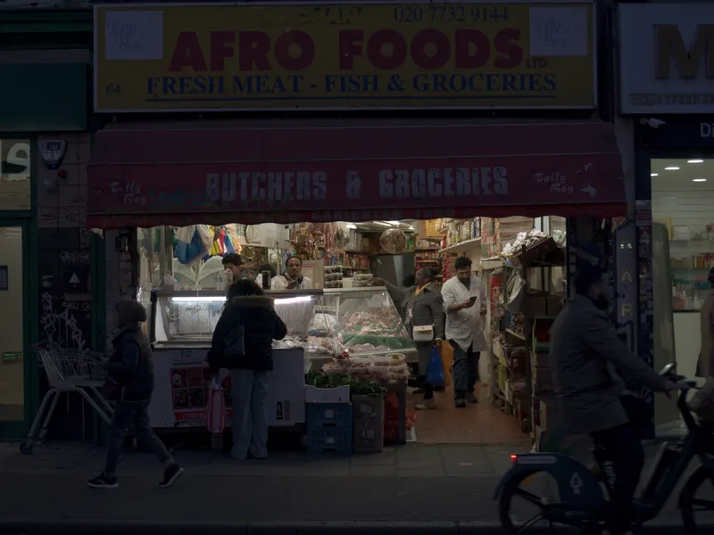 A butchers and grocers lit up at night, a cyclist passing across the frame