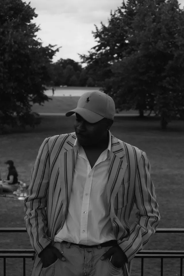 A man in a striped shirt and cap standing in a park, black and white