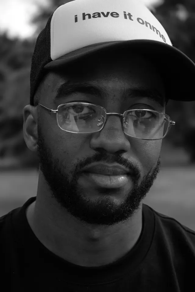 A close black and white portrait of a man in glasses and a slogan cap
