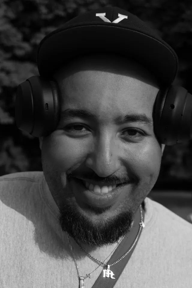 A man smiling wide with headphones around his neck, black and white