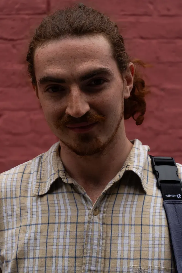 A man with his curly hair tied back, against a painted brick wall