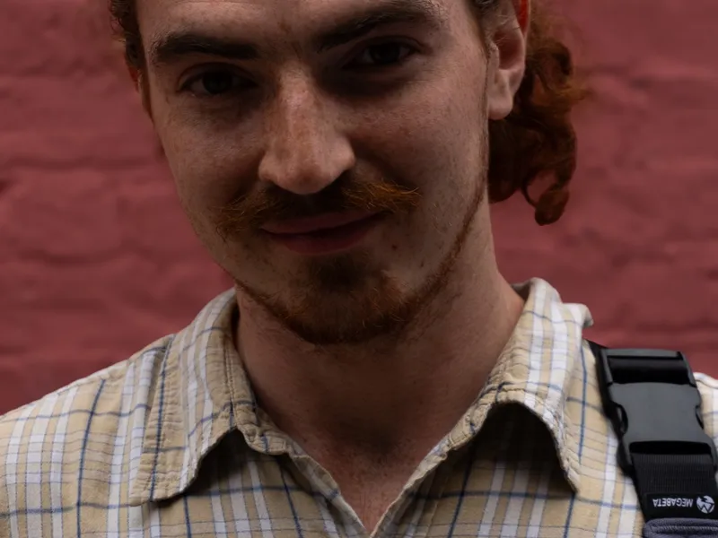 A man with his curly hair tied back, against a painted brick wall
