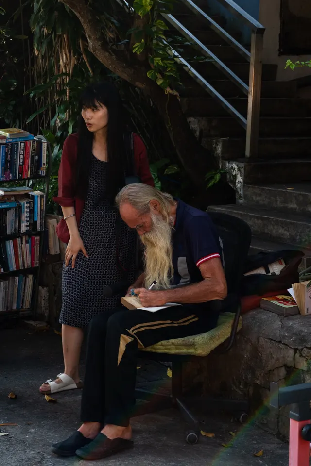 A woman standing beside an older man writing at a roadside bookstall