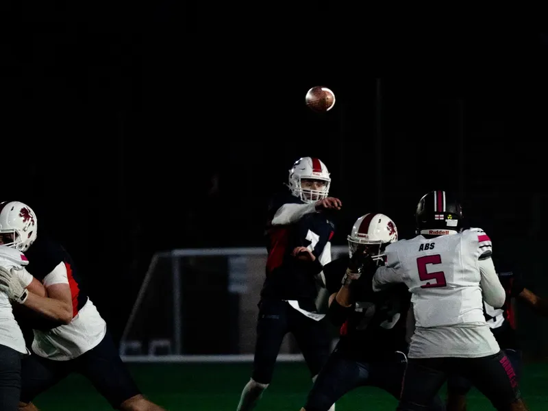 A pass thrown under floodlights, players converging beneath the ball