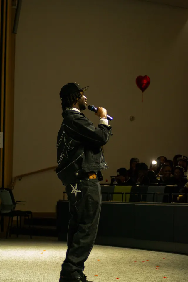 A performer in dark workwear on stage, a red heart balloon drifting behind them