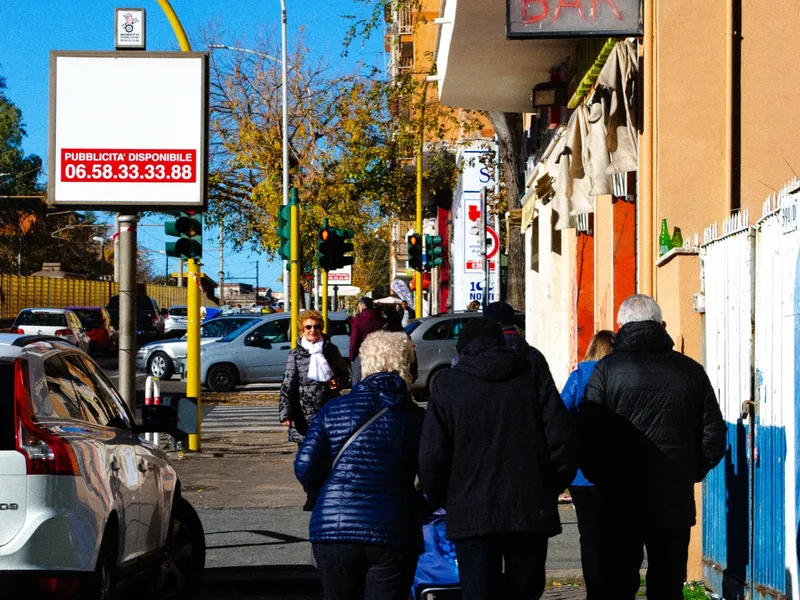 People walking a sunlit street past parked cars and shopfronts