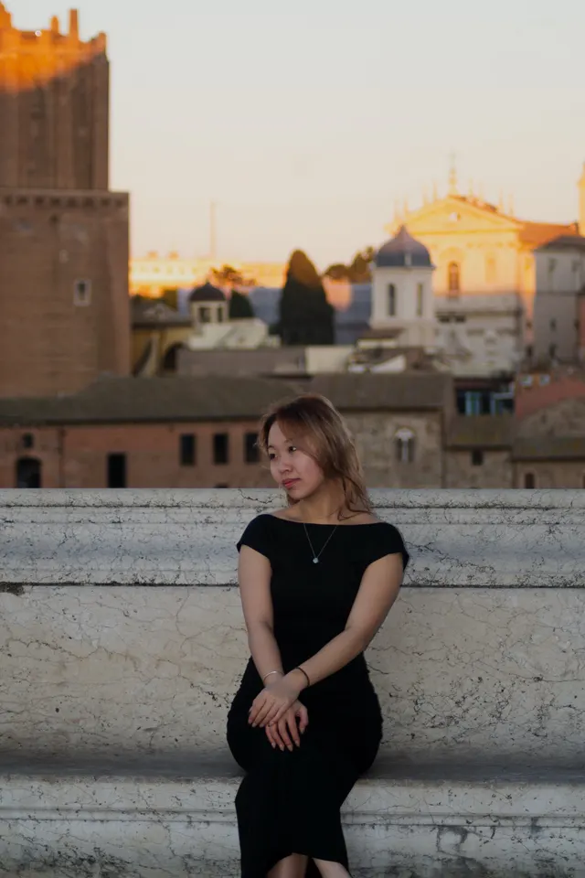 A woman in a black dress seated on marble steps, Rome's rooftops behind her