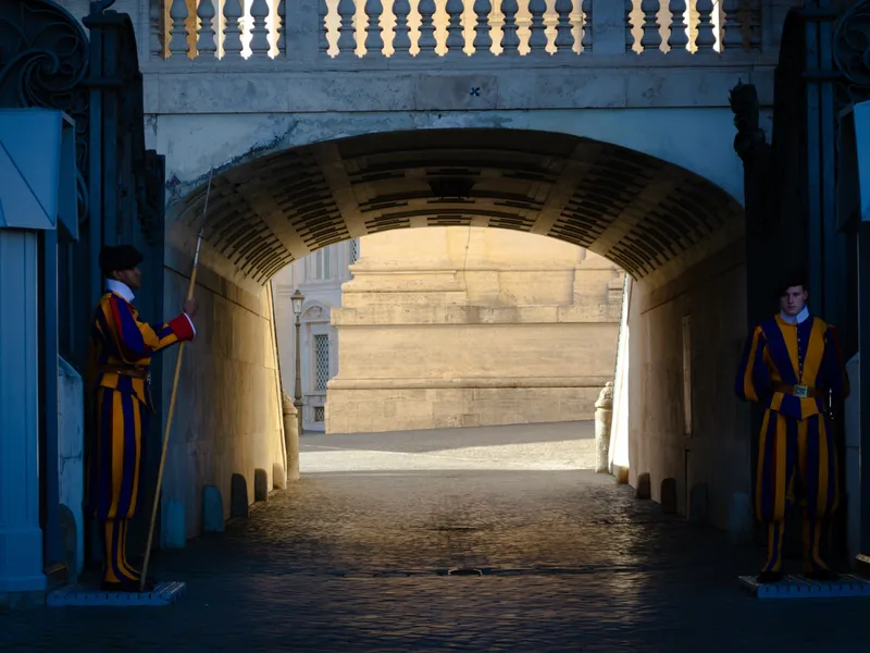 Two Swiss Guards in striped ceremonial uniform standing either side of a sunlit stone archway